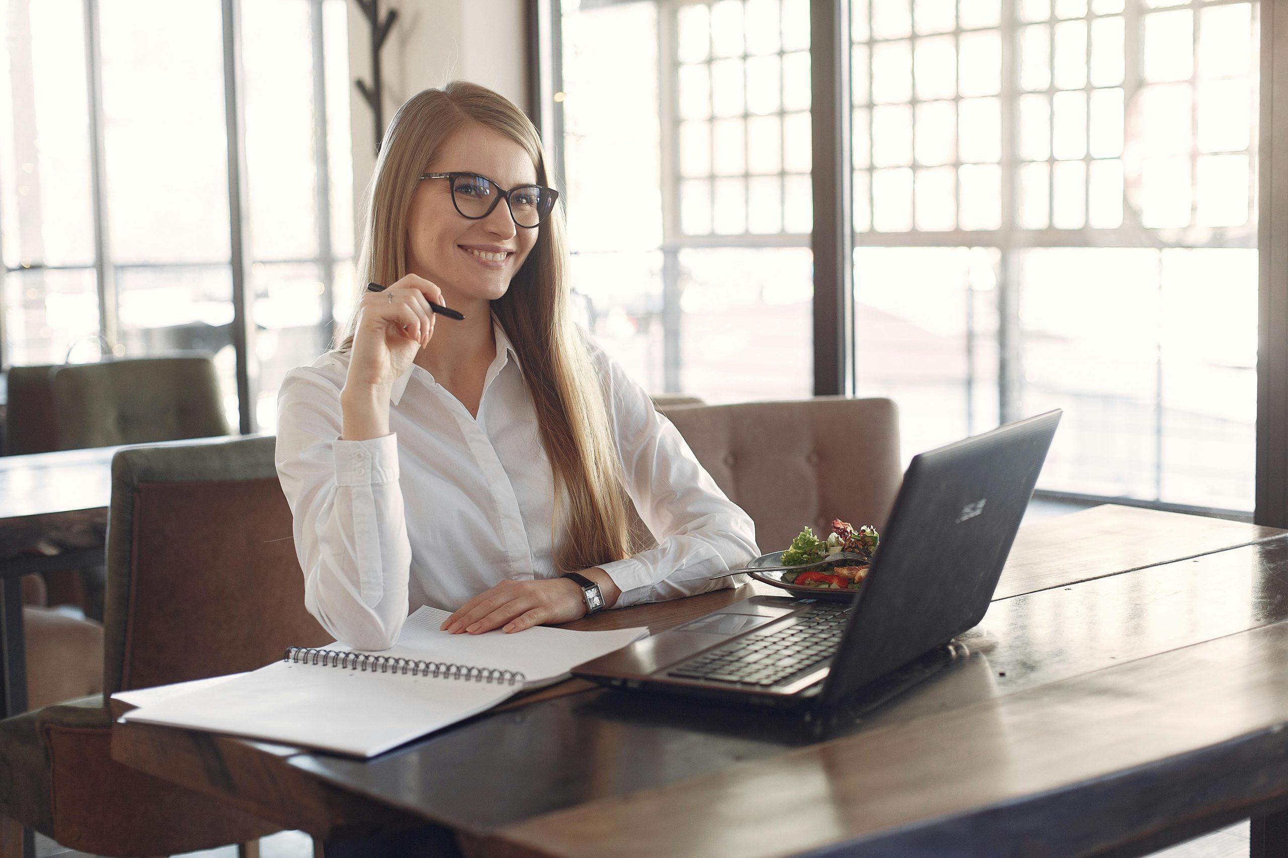 Positive smiling female entrepreneur in formal clothes and eyeglasses sitting at table with laptop and planner holding pen in hand while looking away during lunch break with salad in cafe